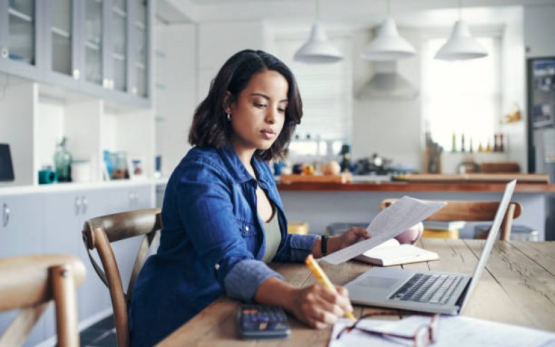 Shot of a young woman using a laptop and  going through paperwork while working from home